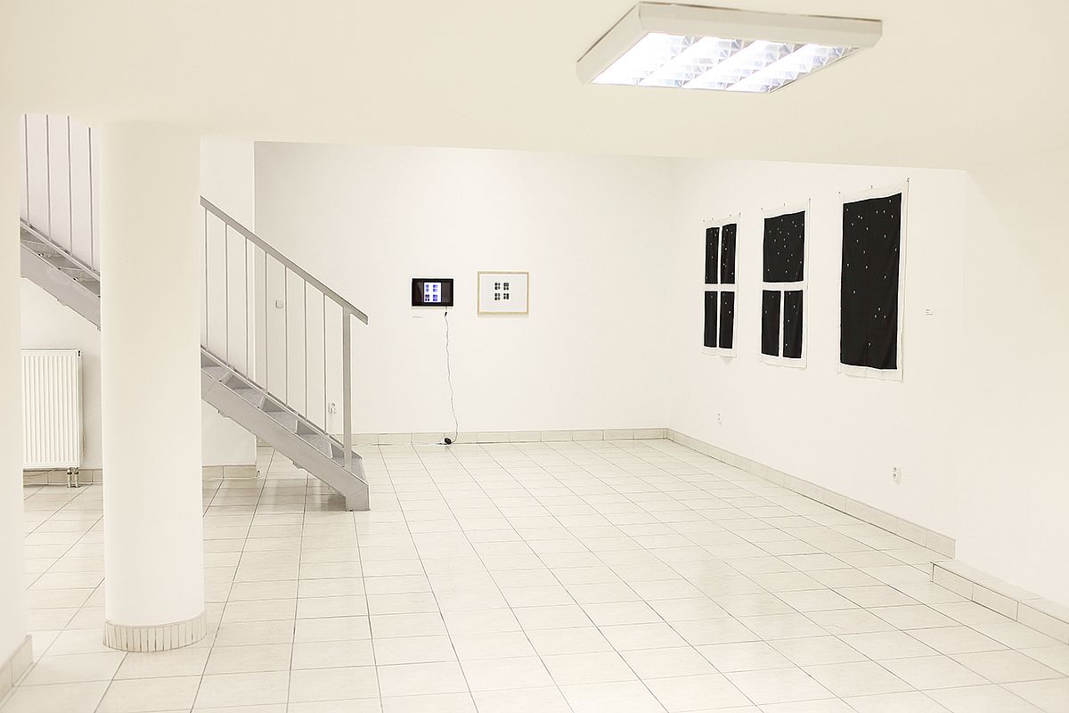 Zdjęcie wnętrza galerii. Ściany, sufit i podłoga w kolorze białym, po lewej stronie widoczne schody. Na ścianach zawieszone niewielkie obrazy.